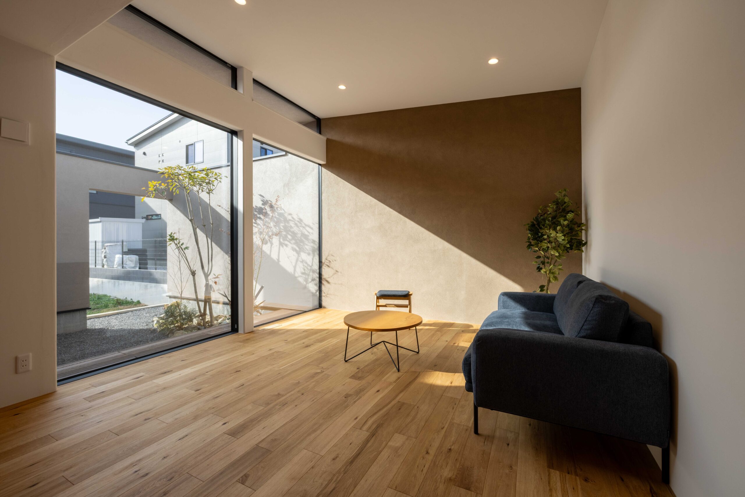 中庭を正面に望む大きなピクチャーウインドーを据えたワンフロアのリビング空間。大開口から差し込む潤沢な光と植栽が相まって、アクセントウォールに陰影を落とす。家族がくつろぐ目線に合わせた設計は、住宅地であることを忘れるほどの安らぎをもたらす。